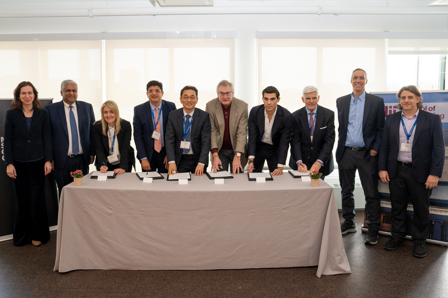10 people stand shoulder-to-shoulder behind a table. Six are bending over to sign a paper in front of each of them.
