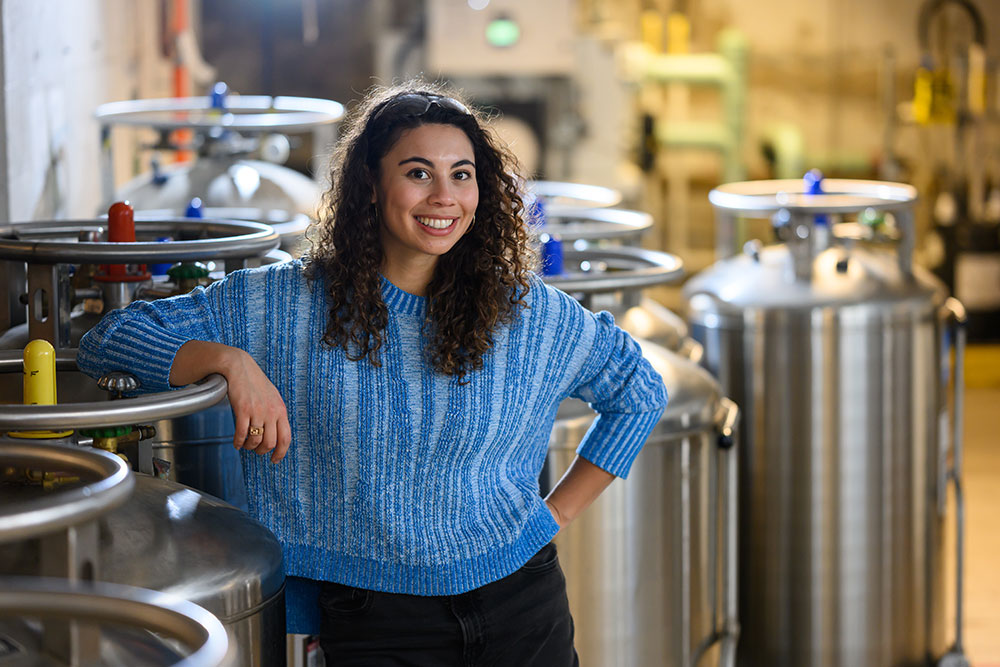 Graduate student, Zoe Fisher, in a lab sapce with stainless steel dewars/tanks in the background