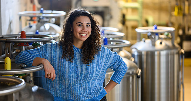 Graduate  student, Zoe Fisher, in a lab sapce with stainless steel dewars/tanks in the background